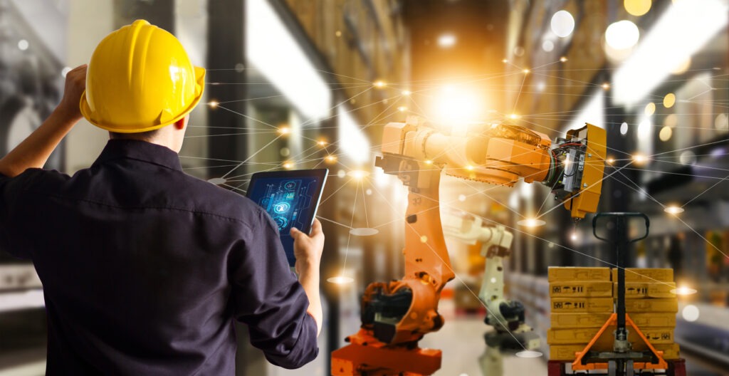 A worker with a yellow hard hat uses a tablet to monitor an automated robotic arm in a factory, symbolizing industrial automation and technology.
